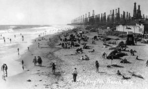 Angelenos play below derricks at Huntington Beach pier, c. 1930-40. In the early decades of the twentieth century, LA residents accepted ground subsidence and small earthquakes as the cost of producing oil. But discontent began to rumble in the late 1930s. Rapid exploitation of Wilmington, one of the largest oil fields in California, and the Huntington Beach field caused Long Beach harbor to sink, buckling railway lines and forcing the city to build higher piers. Credit: Orange County Archives